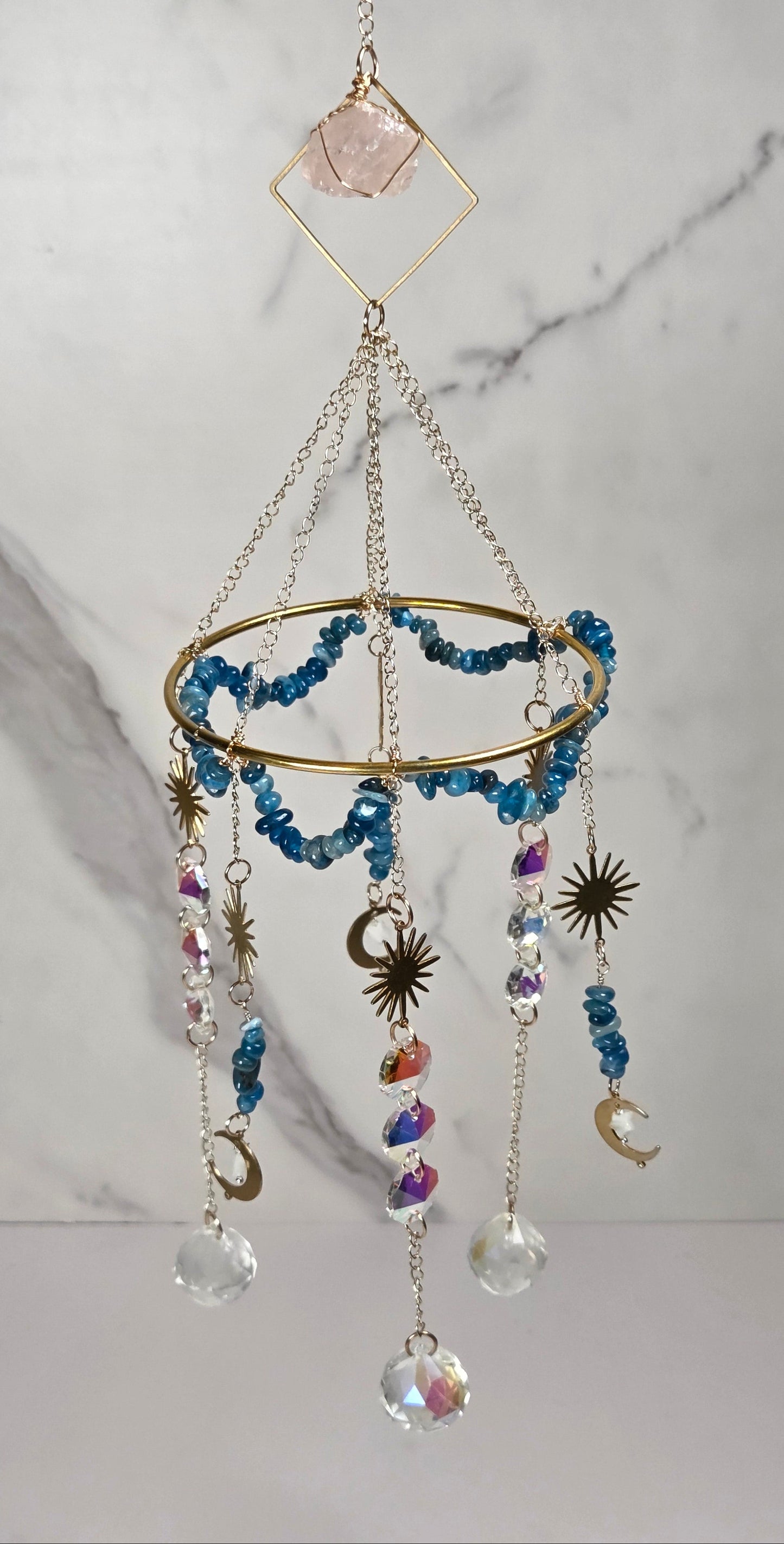 A bohemian-style hanging mobile ornament featuring a gold ring, blue beads, clear crystal drops, and celestial charms, suspended against a white marble background.