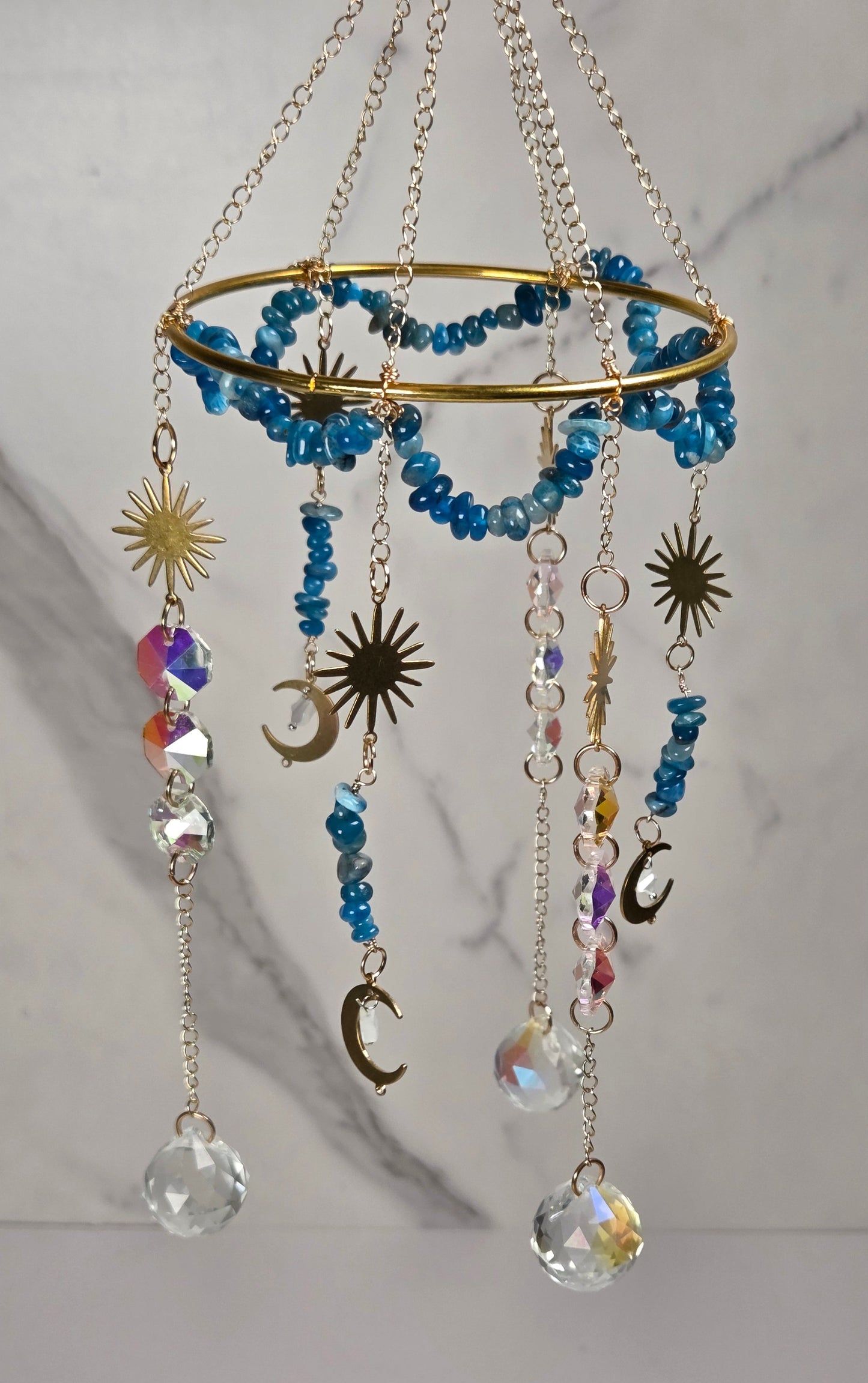 A hanging decorative mobile with a gold-tone circular frame, blue beaded rings, metallic sun and moon charms, and clear faceted crystal drops against a light marble-patterned background.