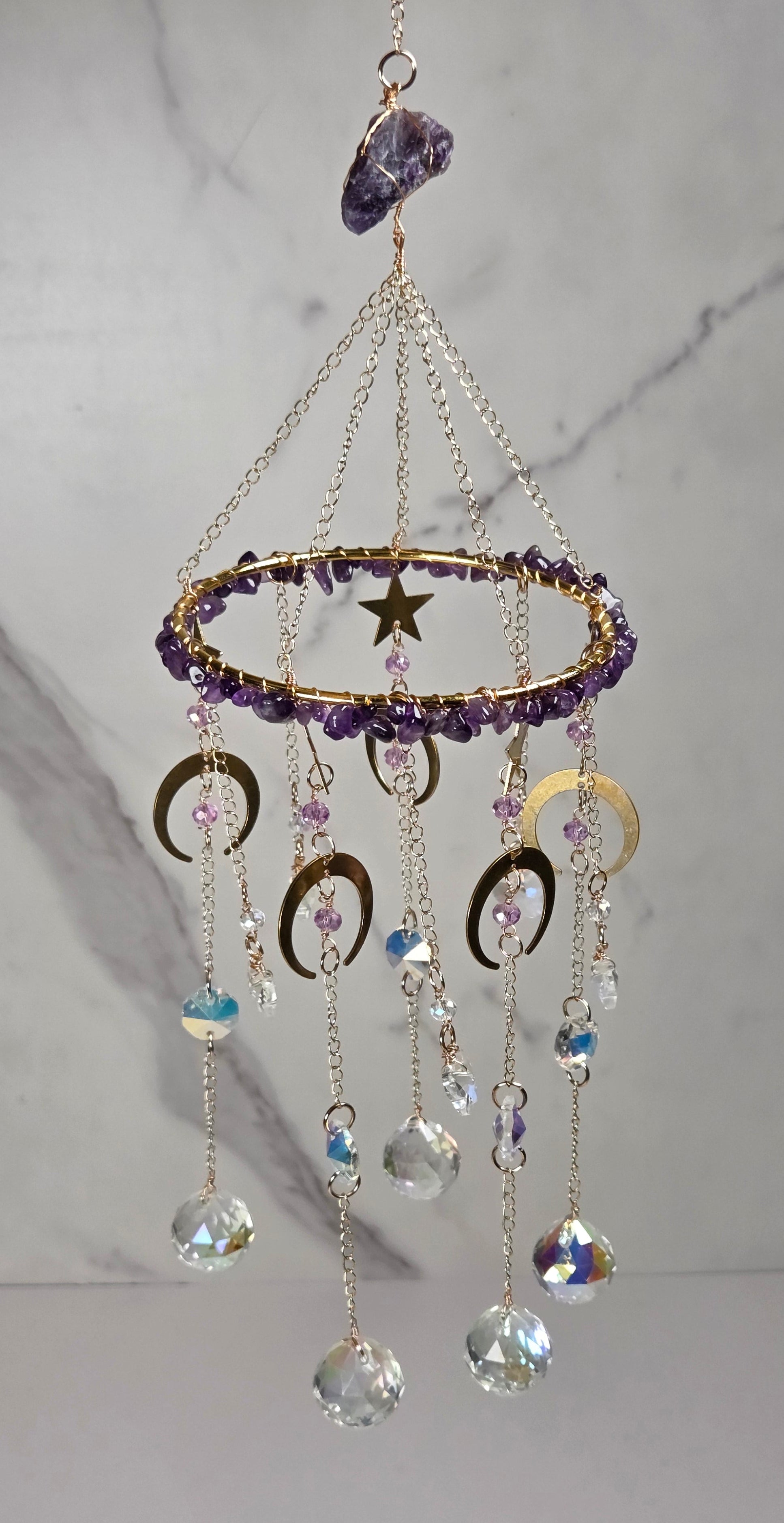 Amethyst celestial mobile hanging against a light gray marble-patterned background. The mobile features a gold-toned ring with purple amethyst beads, a gold star, and dangling gold crescent moon and sun disc charms with clear and iridescent crystal beads.