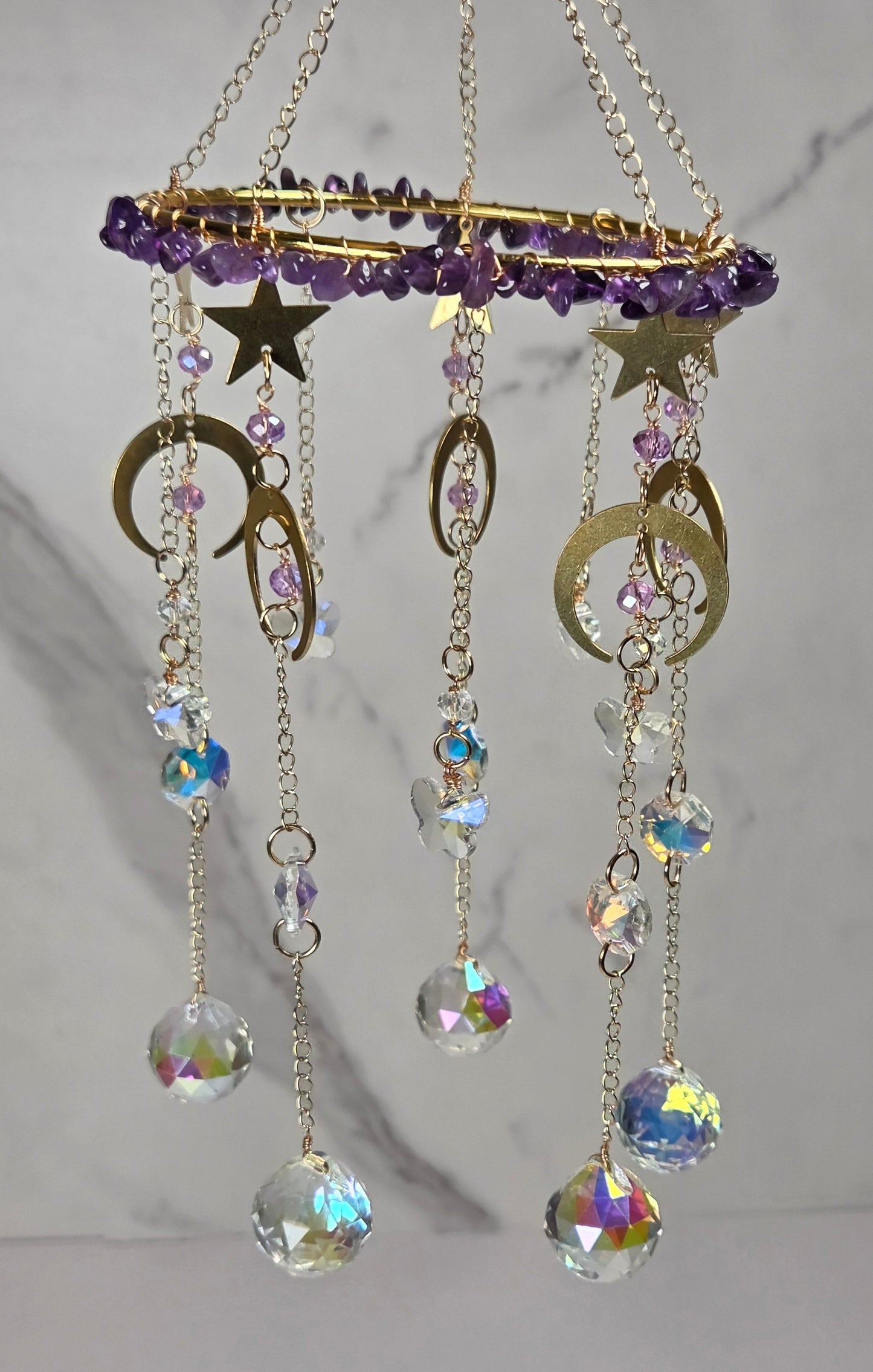 A celestial-themed mobile hanging ornament with gold-toned chains, purple crystal beads, star and moon shapes, and dangling iridescent faceted beads against a light marble-patterned background.