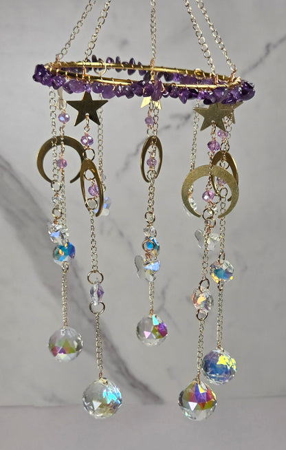 A celestial-themed mobile hanging ornament with gold-toned chains, purple crystal beads, star and moon shapes, and dangling iridescent faceted beads against a light marble-patterned background.