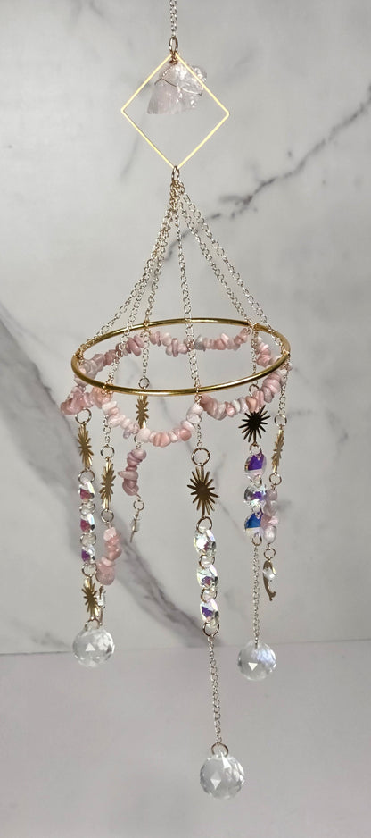 A bohemian-style hanging mobile with a gold-toned circular frame, pink faceted beads, clear crystal drops, and small star and moon charms, suspended against a white marble-patterned background.