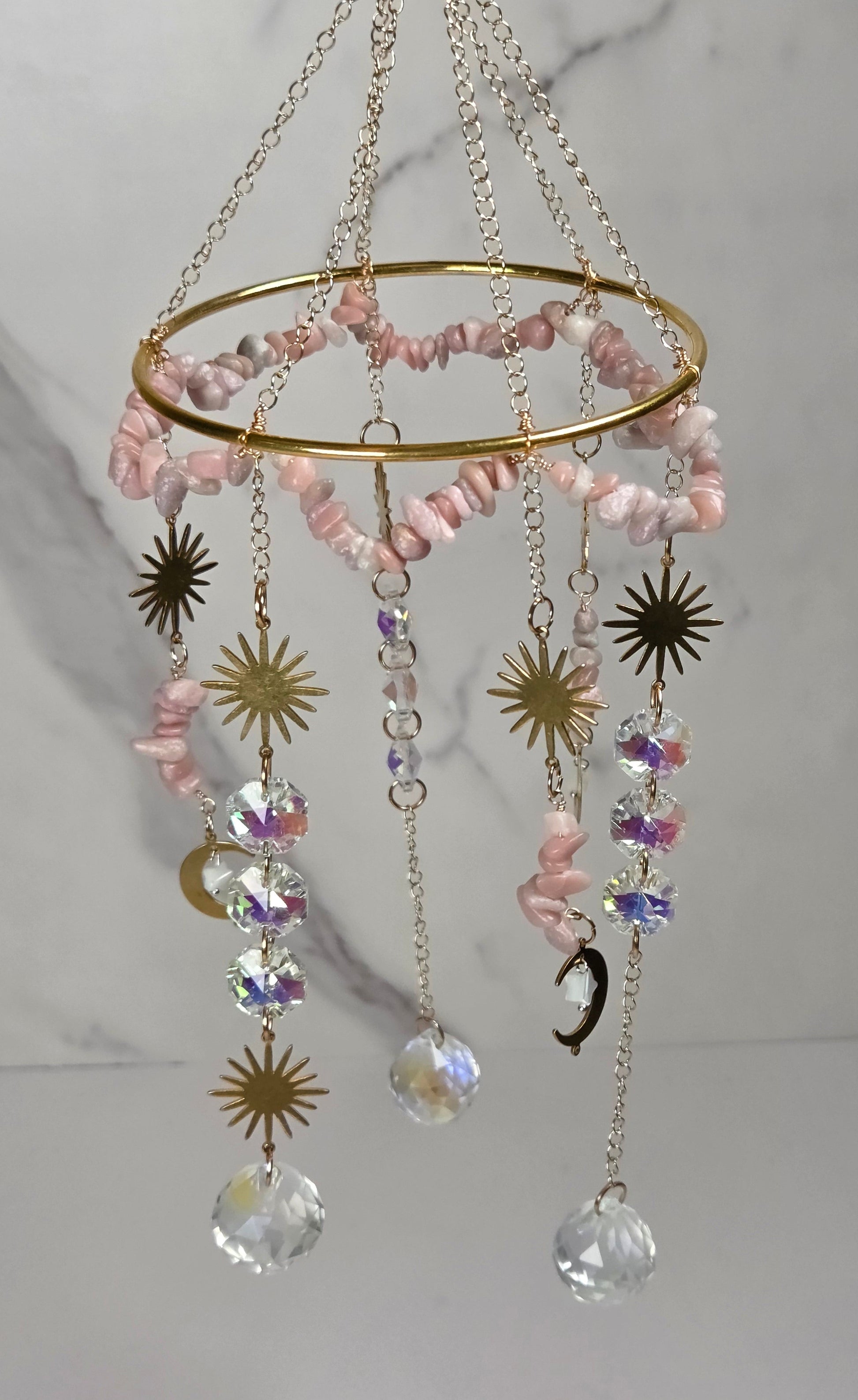 A gold-toned hanging ornament with a circular ring, pink stone beads, dangling crystal drops, and metallic sun and moon charms against a light marble-patterned background.
