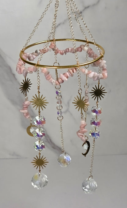 A gold-toned hanging ornament with a circular ring, pink stone beads, dangling crystal drops, and metallic sun and moon charms against a light marble-patterned background.