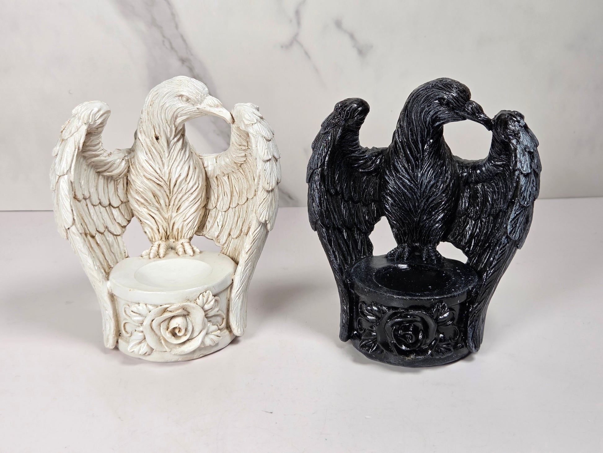 Two decorative eagle figurines with spread wings, each mounted on a circular base with floral detailing. One is off-white/ivory and the other is glossy black, displayed side by side on a light surface against a pale marble-pattern backdrop.