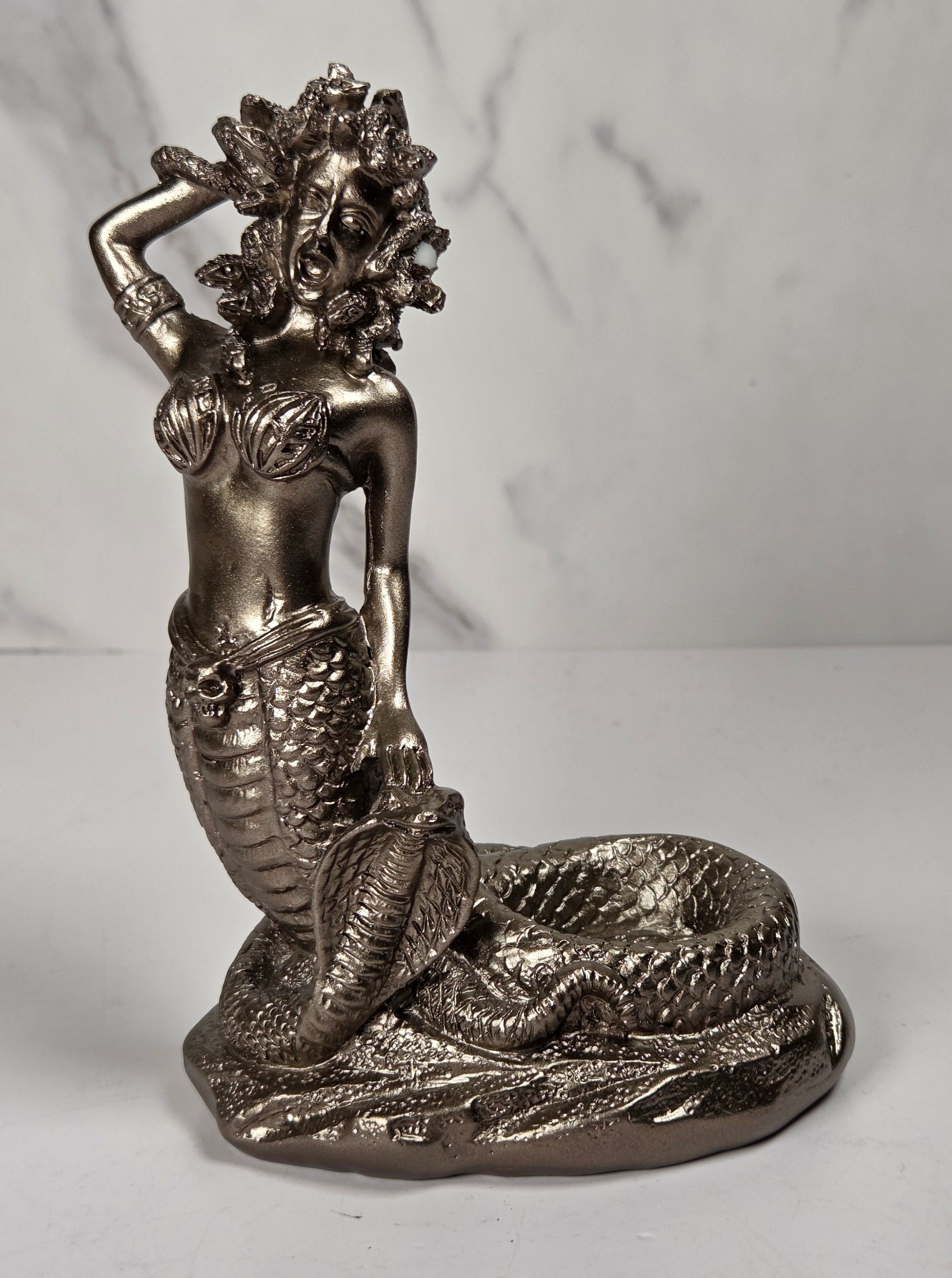 A metallic silver mermaid figurine sitting on a rock base next to a snake, set against a white marble-patterned background.