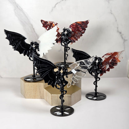 Four decorative winged fairy figurines displayed on black metal stands, arranged on light wood hexagonal blocks against a white marble-patterned background.
