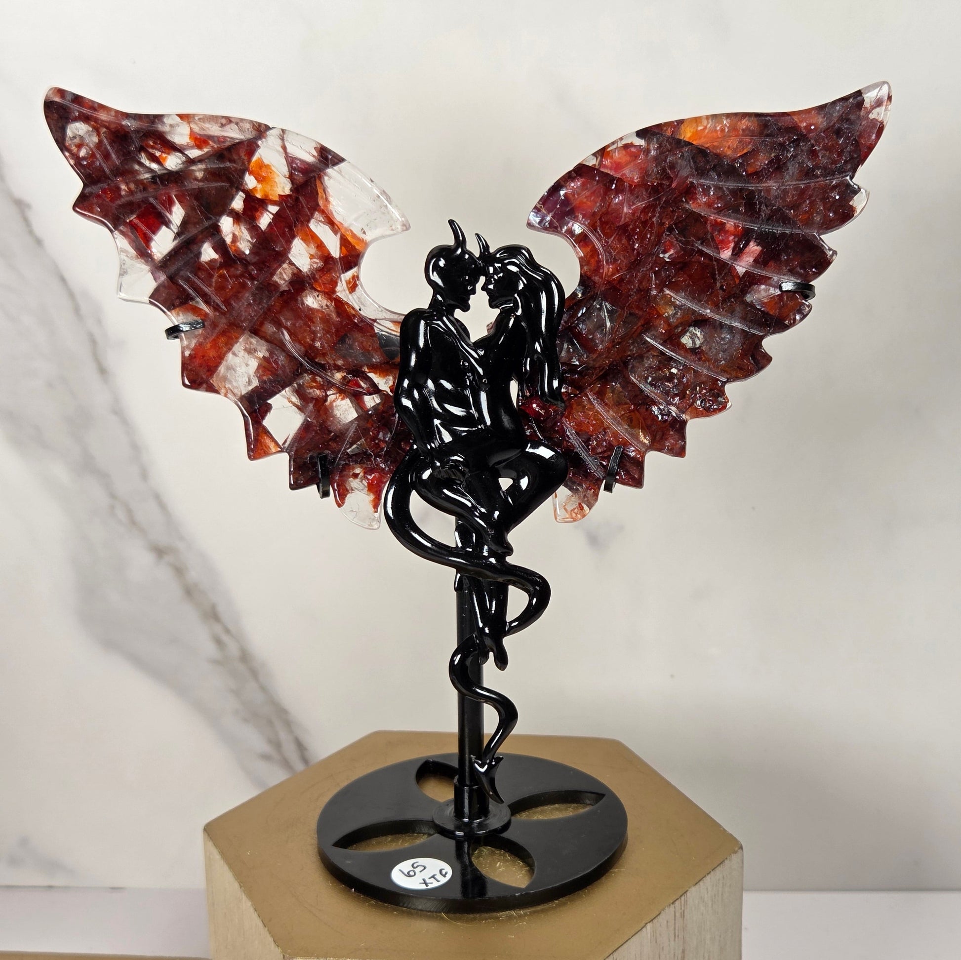 A decorative winged sculpture featuring translucent reddish-brown and amber wings on a black metal figure mounted on a round black base, displayed on a light wood block against a white marble-patterned background.