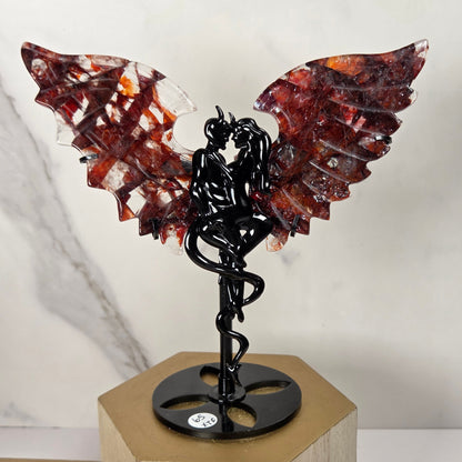 A decorative winged sculpture featuring translucent reddish-brown and amber wings on a black metal figure mounted on a round black base, displayed on a light wood block against a white marble-patterned background.