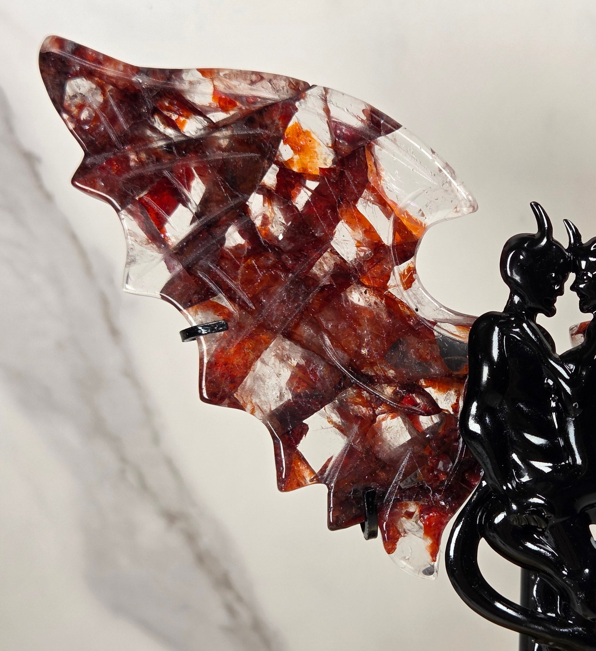 A decorative winged sculpture featuring a glossy black intertwined figure on a stand, with translucent wings containing red, amber, and clear marbled patterns, set against a light marble-patterned background.