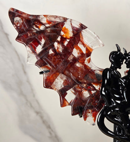 A decorative winged sculpture featuring a glossy black intertwined figure on a stand, with translucent wings containing red, amber, and clear marbled patterns, set against a light marble-patterned background.