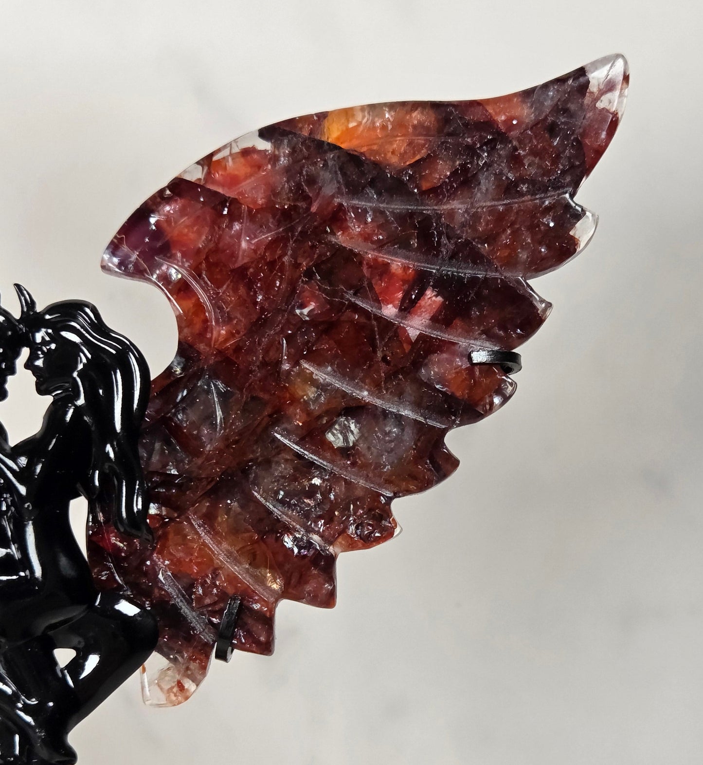 A decorative winged figurine sculpture with a glossy black twisted wire devil-like figure and layered translucent wings in deep red, burgundy, and amber tones, displayed against a light neutral background.