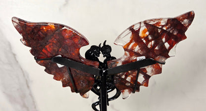 A decorative winged sculpture or ornament with two translucent reddish-brown wings and a black central figure and support, photographed against a light marble-like background.