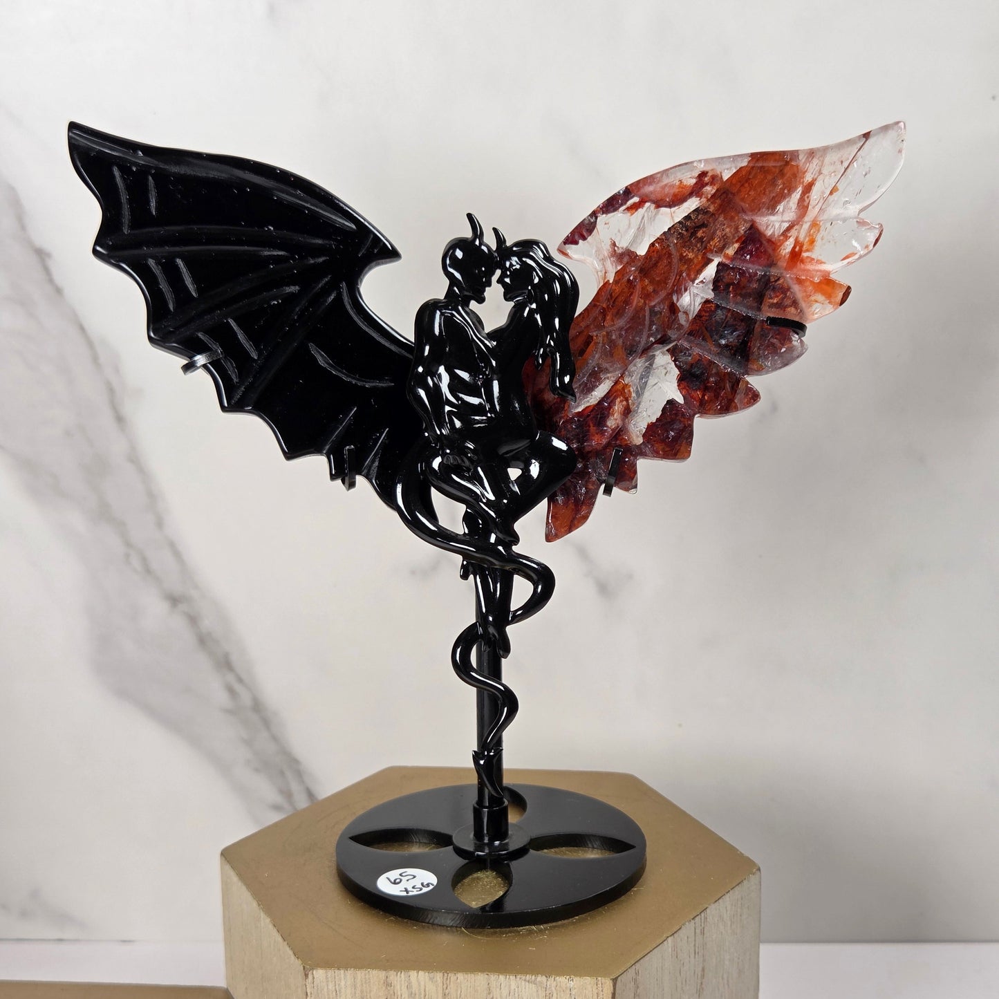 A decorative metal sculpture of a bat-like figure with one black wing and a translucent reddish-brown crystal wing, mounted on a black circular base atop a light wood pedestal against a white marble-patterned background.