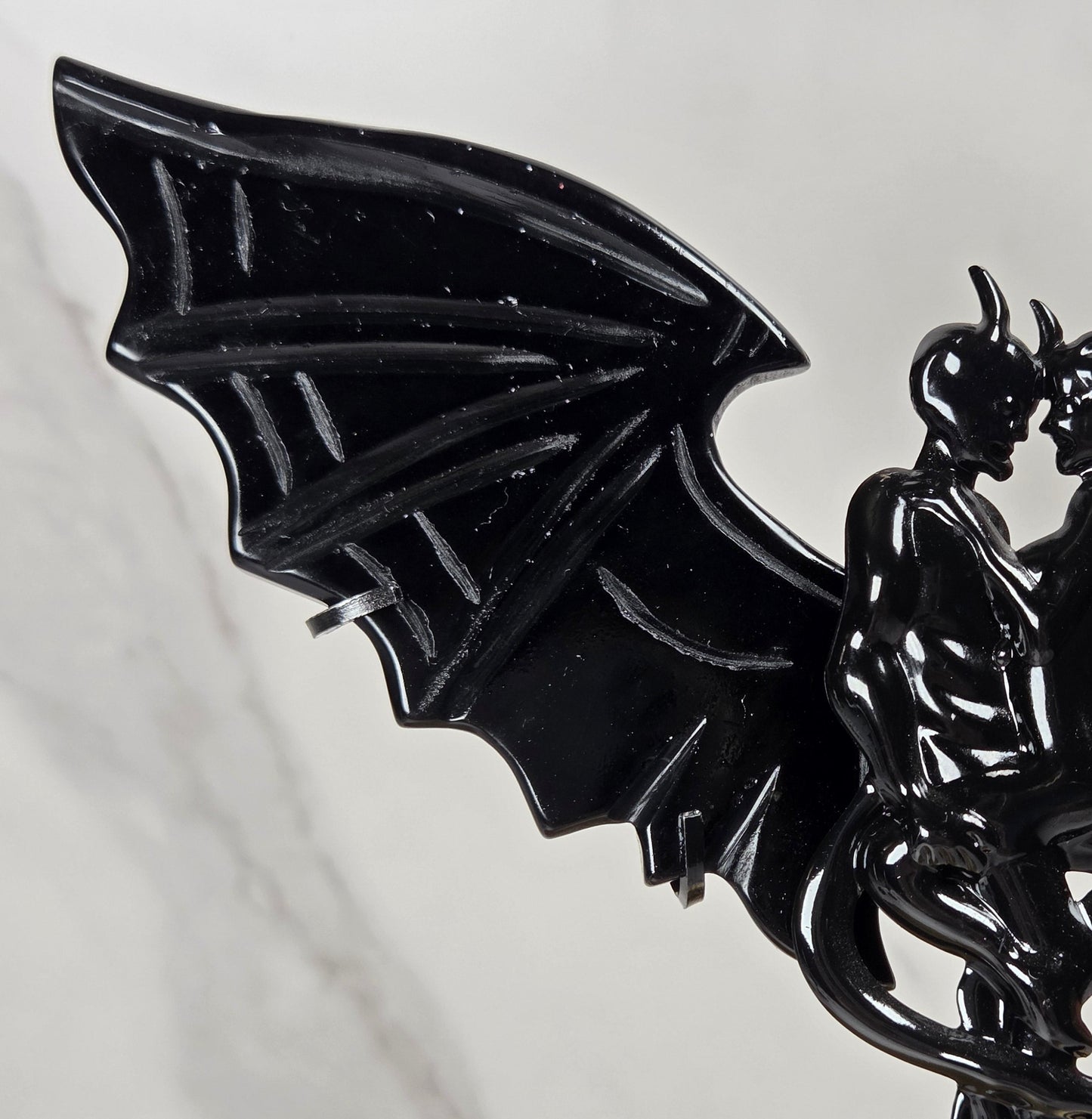 A black gothic figurine of a kissing couple on a staff, featuring a large bat wing on the left and a red crystal cluster on the right, displayed on a light marble background.