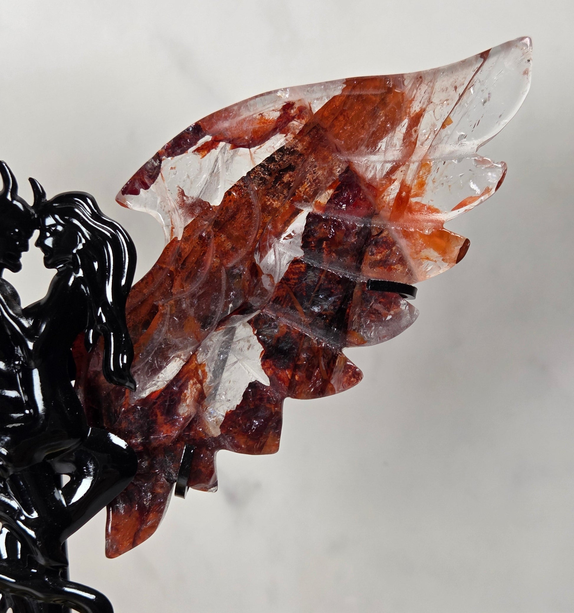 A carved crystal wing sculpture with layered translucent red and brown stone wings mounted on a black metal devil figurine, photographed against a light gray wall.