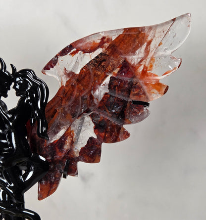 A carved crystal wing sculpture with layered translucent red and brown stone wings mounted on a black metal devil figurine, photographed against a light gray wall.