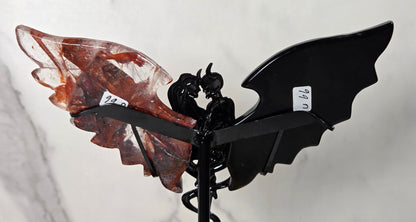 A decorative metal angel wing wind spinner featuring a glossy black bat-wing shaped metal frame and a translucent reddish-brown crystal wing, mounted on a black spiral stake against a light gray marble-patterned background.