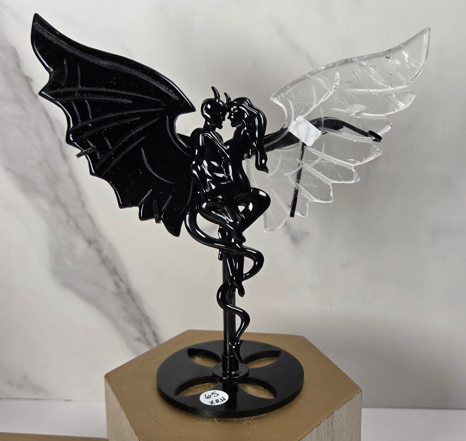 A black metal winged fairy figurine with one black wing and one clear acrylic wing, mounted on a black circular base with a spiral design, displayed on a wooden block against a marble-patterned background.