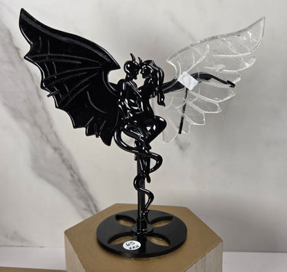 A black metal winged fairy figurine with one black wing and one clear acrylic wing, mounted on a black circular base with a spiral design, displayed on a wooden block against a marble-patterned background.