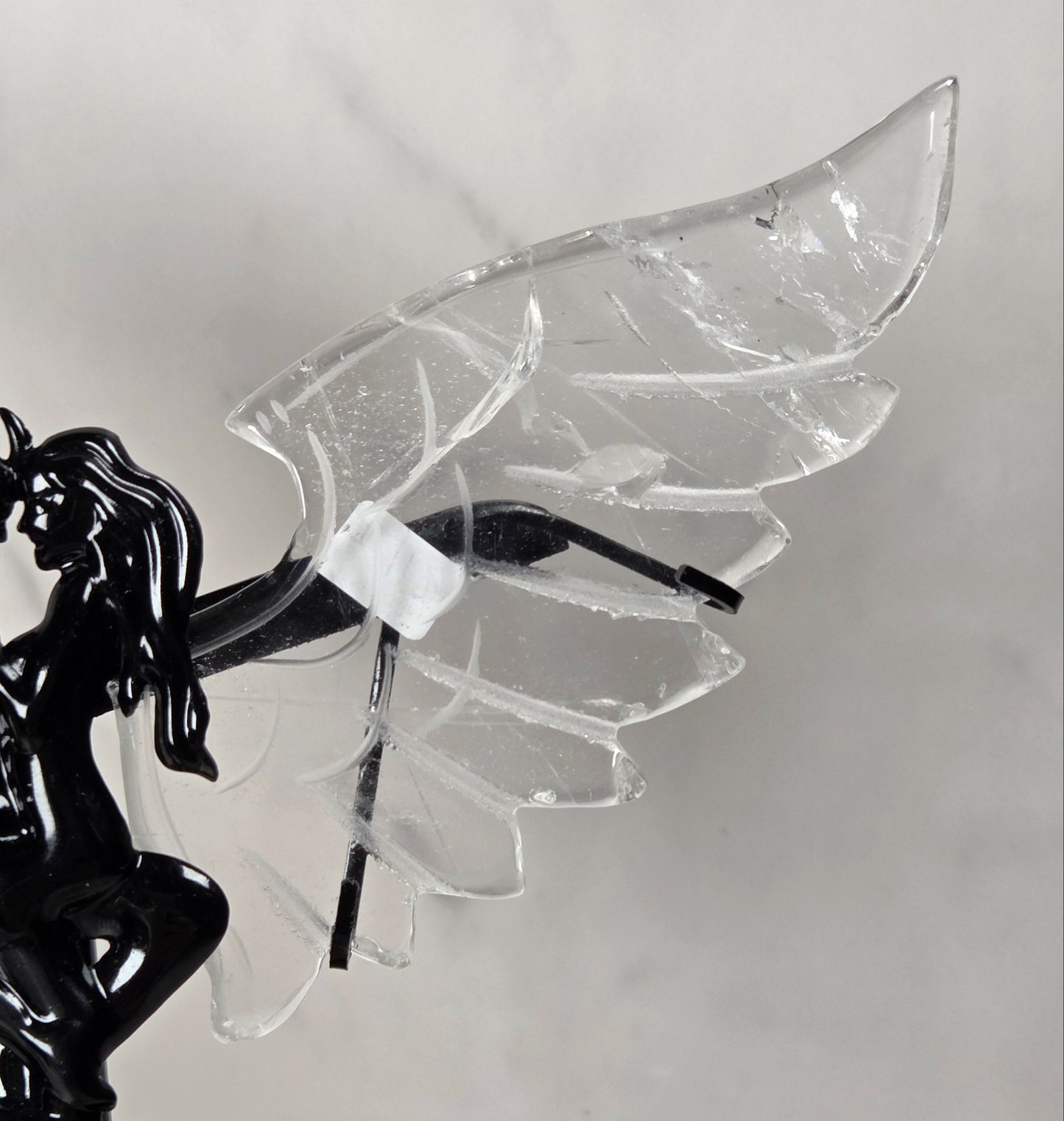 A black metal winged fairy figurine with a glossy finish, featuring a stylized couple embracing on the left and a large translucent clear wing on the right, shown against a light gray background.