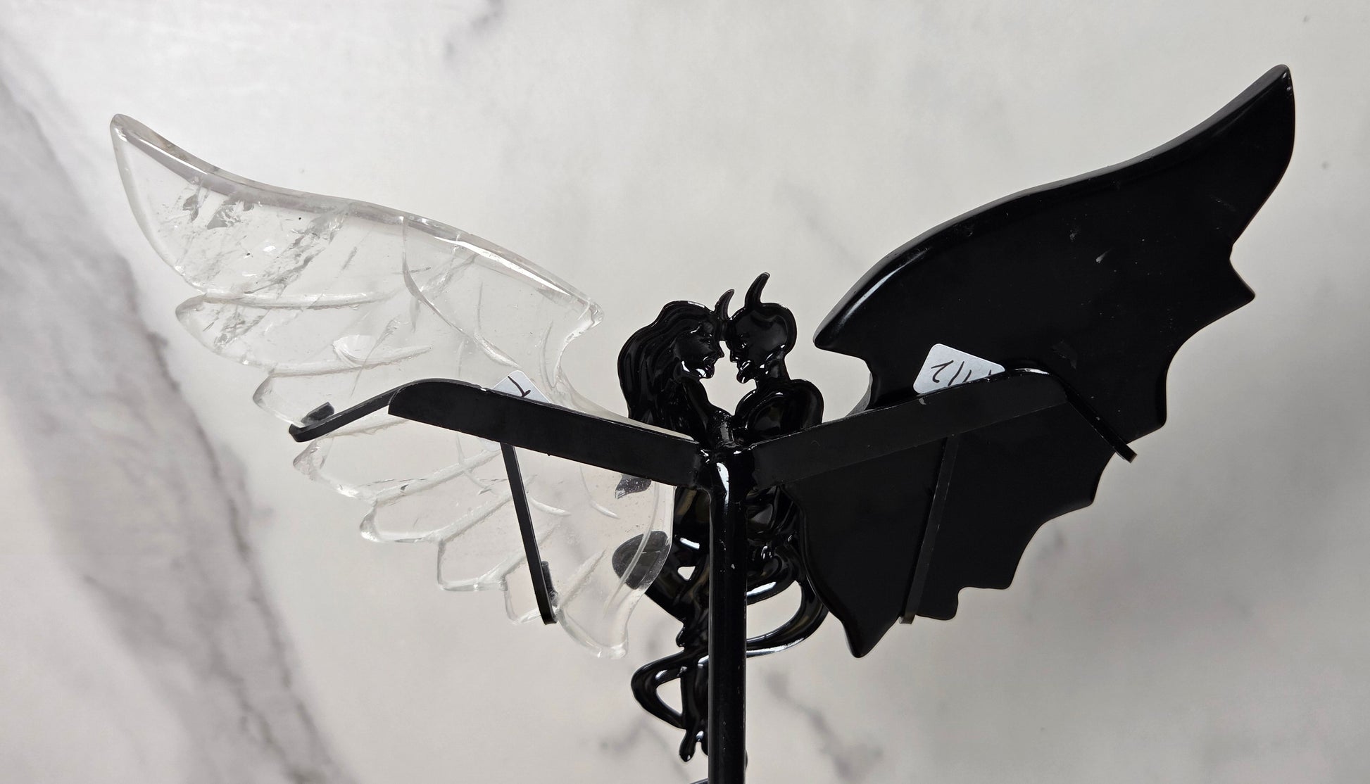 A gothic-style metal garden stake featuring a black winged figure with one transparent clear wing and one solid black wing, set against a light gray marble-patterned background.