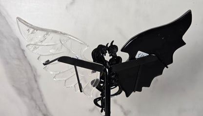 A gothic-style metal garden stake featuring a black winged figure with one transparent clear wing and one solid black wing, set against a light gray marble-patterned background.