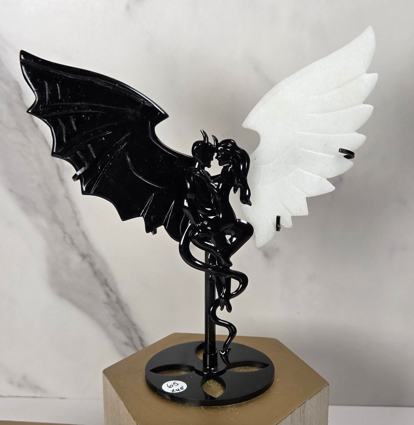 A decorative winged figure sculpture on a stand, featuring a black bat-like wing on the left, a white feathered wing on the right, and a black central body with a snake motif. The piece is displayed on a gold hexagonal pedestal against a light marble-patterned background.