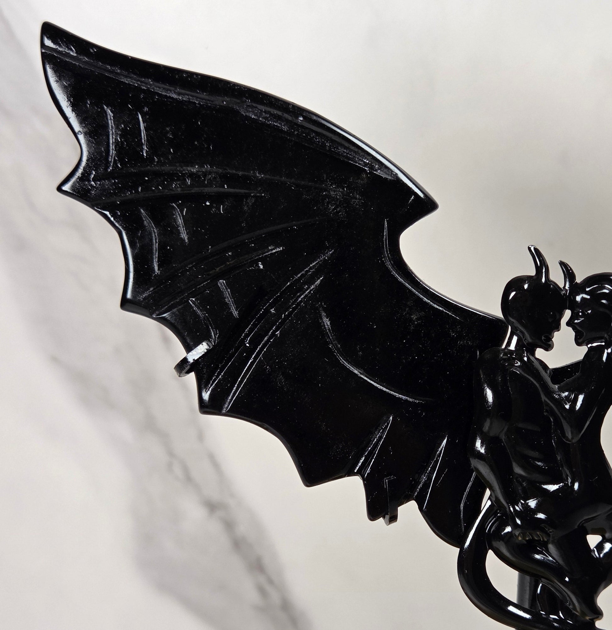 A glossy black bat-winged fantasy figurine or wall decor piece mounted on a thin stand, photographed against a light marble-patterned background.
