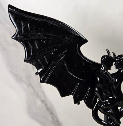 A glossy black bat-winged fantasy figurine or wall decor piece mounted on a thin stand, photographed against a light marble-patterned background.
