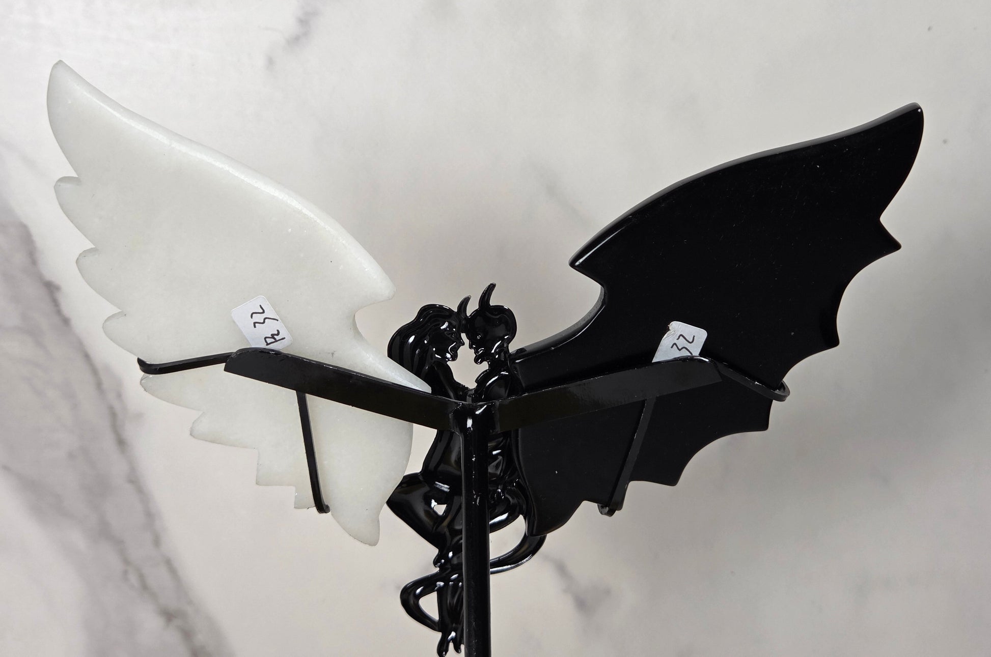 A decorative metal garden stake featuring a black bat-wing design with one glossy black wing and one matte white wing, mounted on a black spiral stake against a light marble-patterned background.