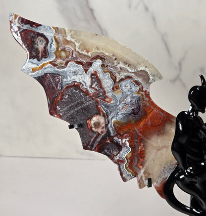 A decorative bat-shaped stone carving or plaque with marbled agate-like patterns in cream, gray, rust red, and dark brown, displayed against a light marble-patterned background next to a glossy black figurine.