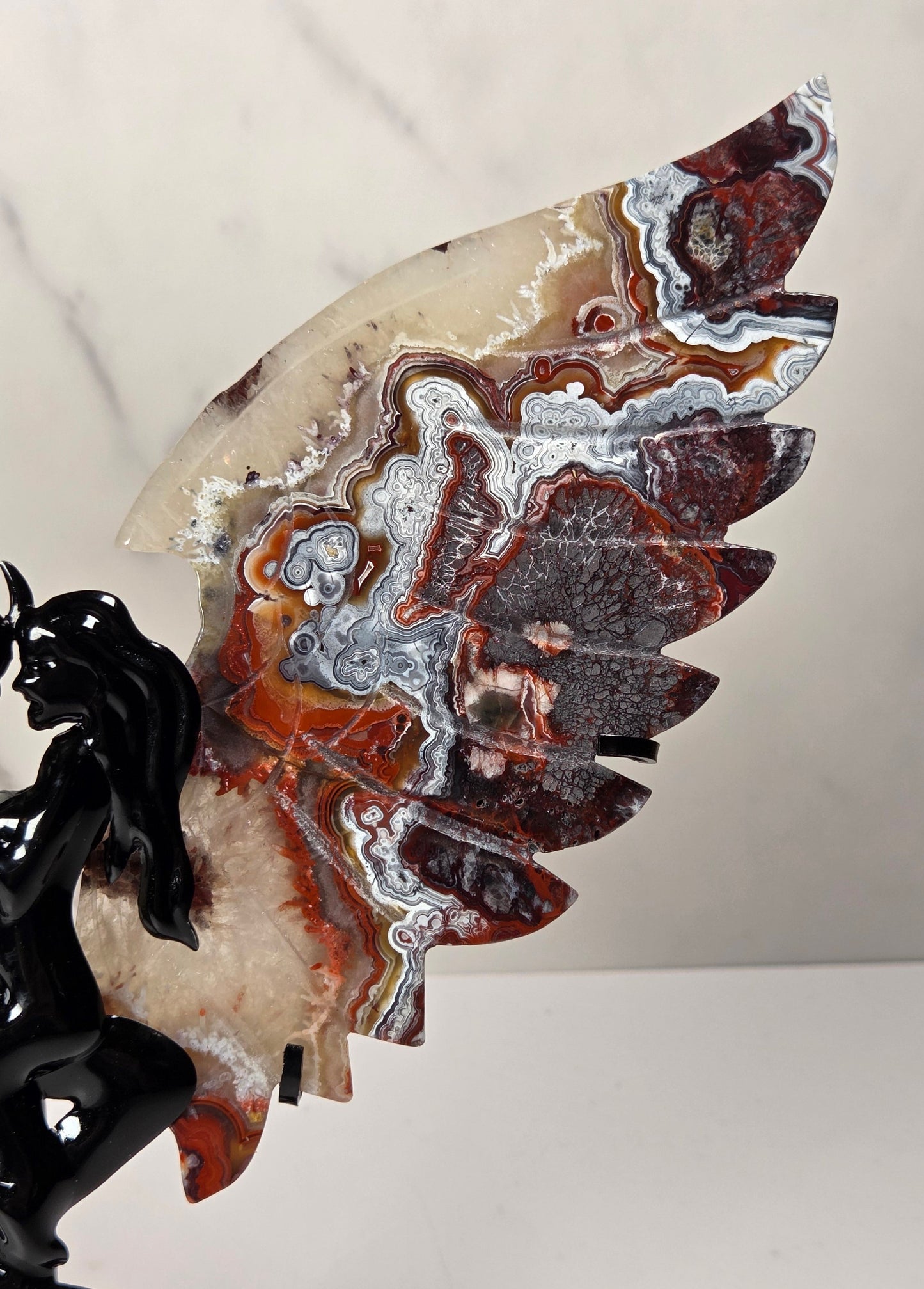 A decorative wall sculpture featuring a polished agate wing with marbled red, brown, cream, and silver patterns, mounted on a glossy black devil figurine, set against a light marble-patterned background.