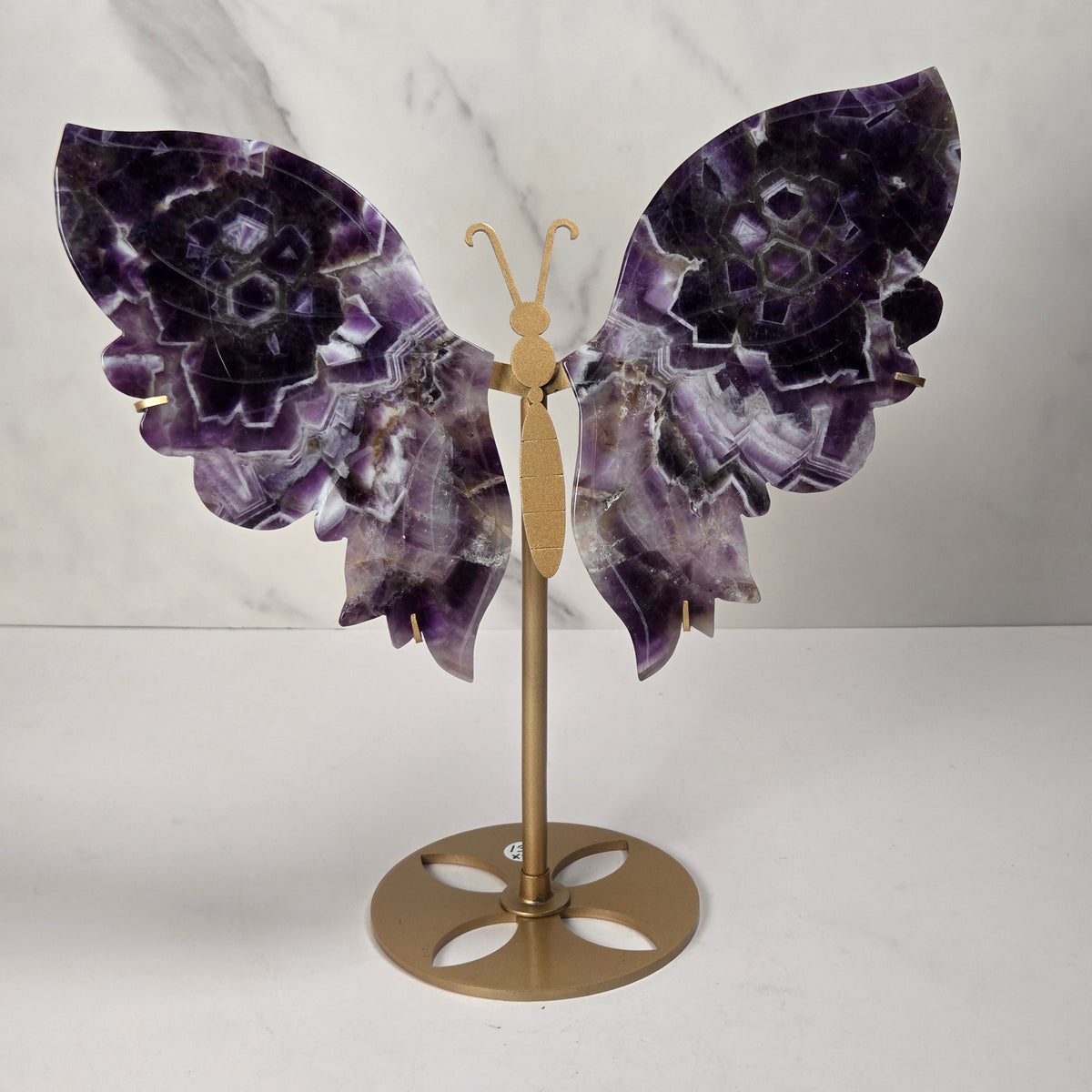 A decorative butterfly wall sculpture featuring purple and white amethyst stone wings mounted on a gold metal stand, set against a white marble-patterned background.