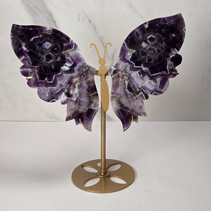 A decorative butterfly wall sculpture featuring purple and white amethyst stone wings mounted on a gold metal stand, set against a white marble-patterned background.