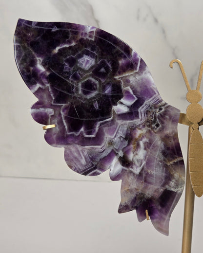 A decorative butterfly sculpture made from purple and white marbled stone wings mounted on a gold-colored metal stand, set against a white marble-patterned background.