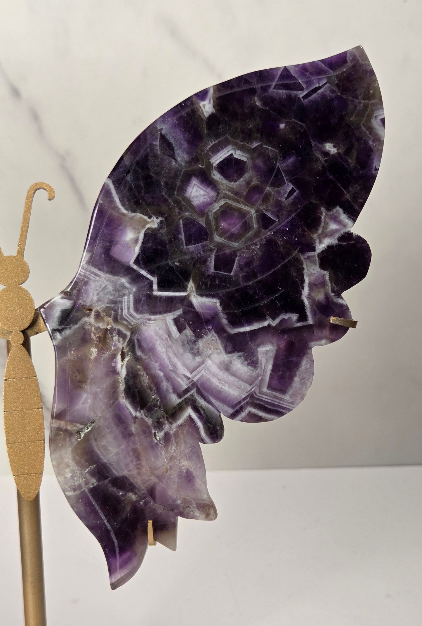 A butterfly-shaped wall decor piece made from polished purple amethyst stone wings, mounted on a small gold-colored metal stand against a light gray marble-patterned background.