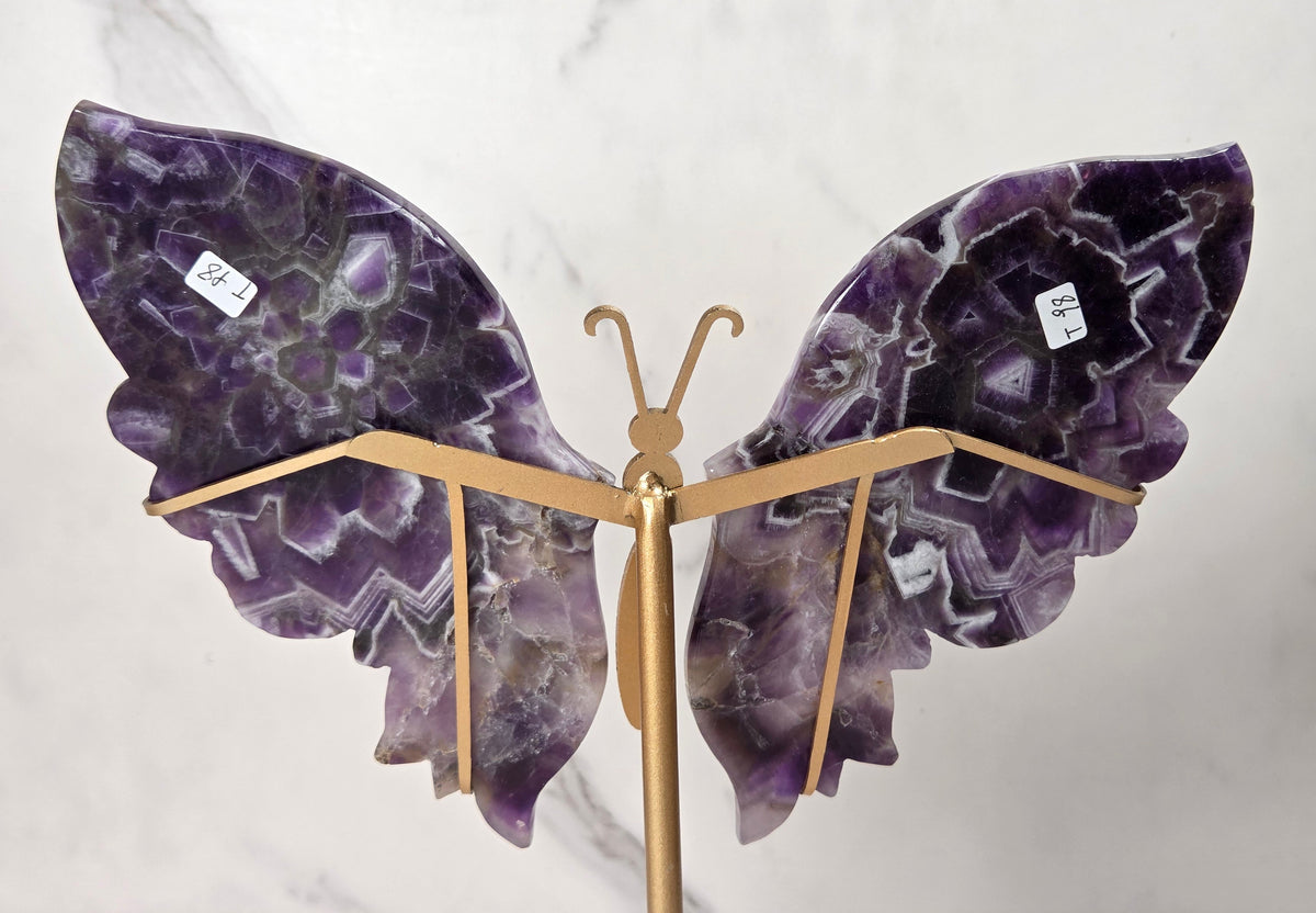 A decorative butterfly sculpture made from polished purple amethyst stone wings mounted on a gold-colored metal stand, set against a white marble background.
