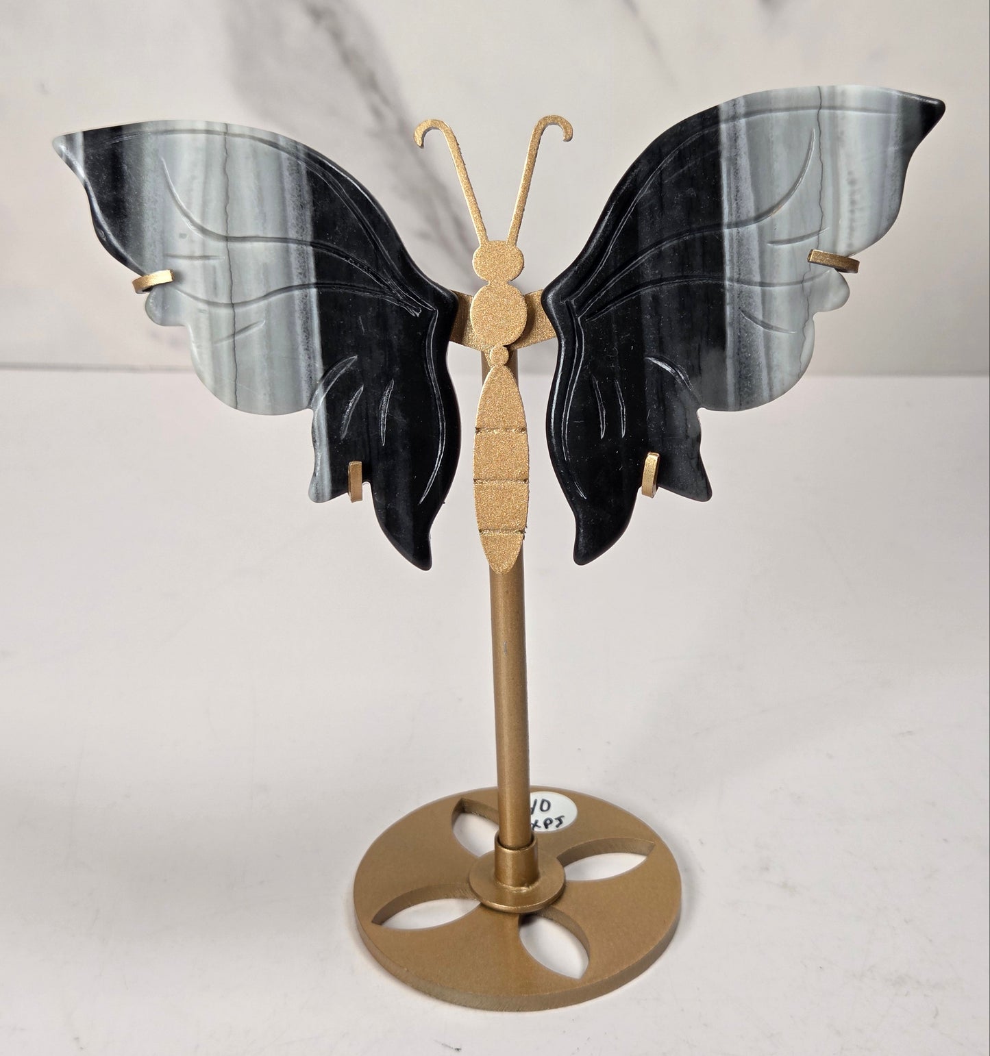 A decorative butterfly-shaped jewelry display stand with black and gray marbled wings on a gold-tone metal stand with a round base, set against a white marble-patterned background.