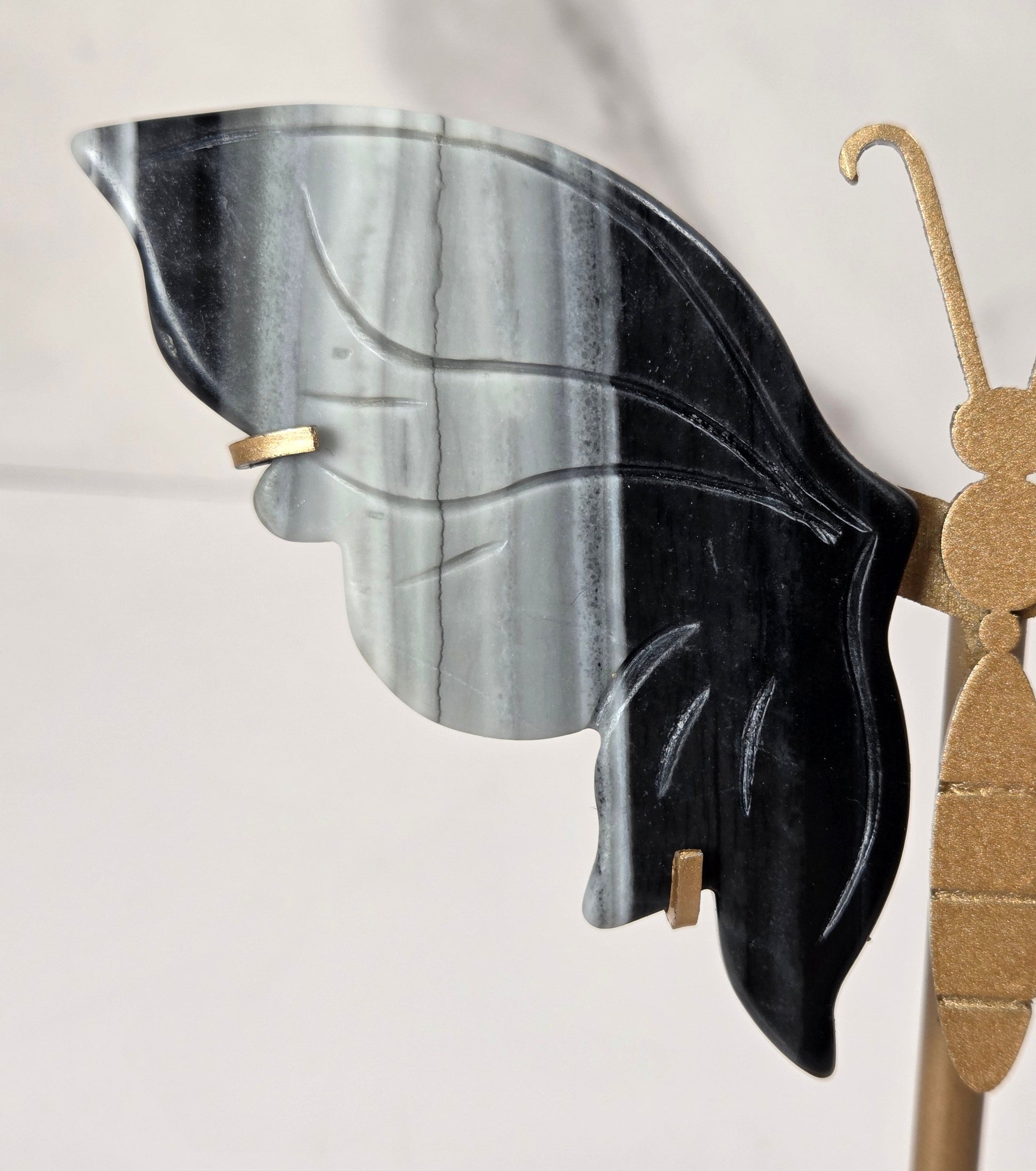 A close-up of a decorative butterfly wall art piece featuring one black and gray marbled wing and one gold-toned wing, mounted on a gold-toned stand against a light marble background.