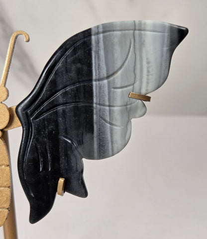 A close-up of a decorative butterfly wing wall piece with one black wing and one gray-and-white marbled wing, mounted on a gold-toned stand against a light marble-patterned background.