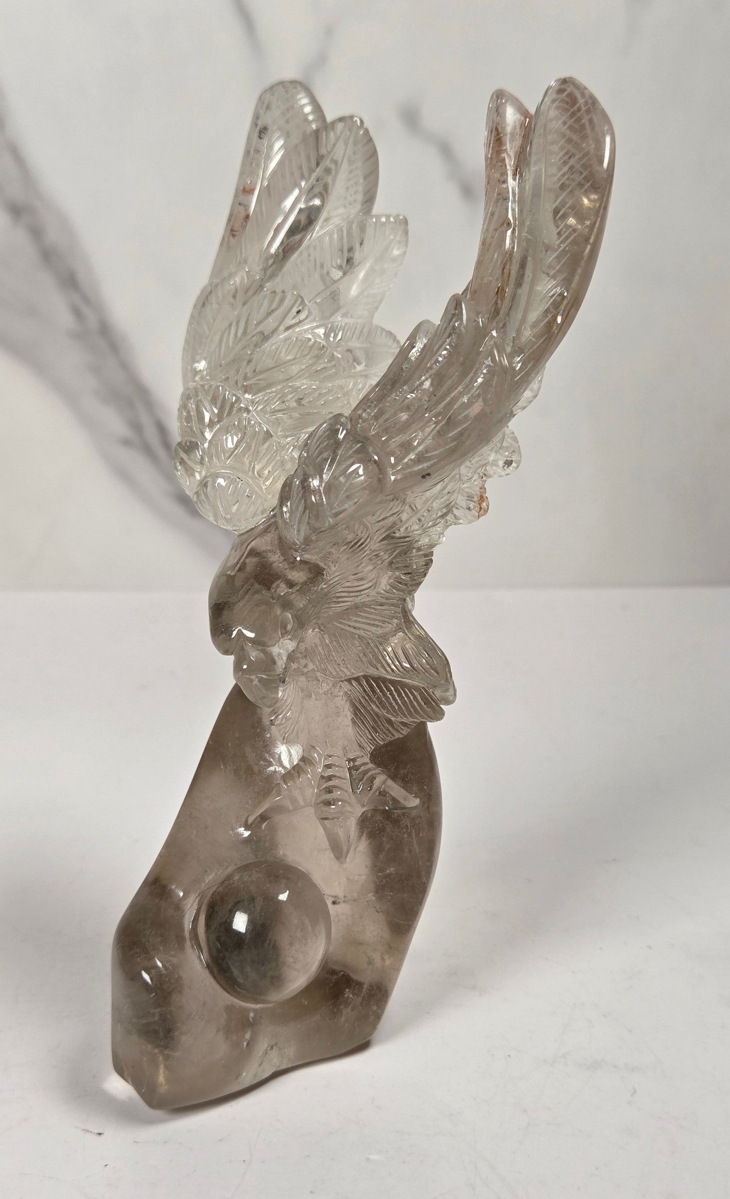 A clear smoky quartz crystal sculpture of a stag head with antlers and a round crystal ball embedded in the base, displayed on a white surface against a light gray marble-patterned background.