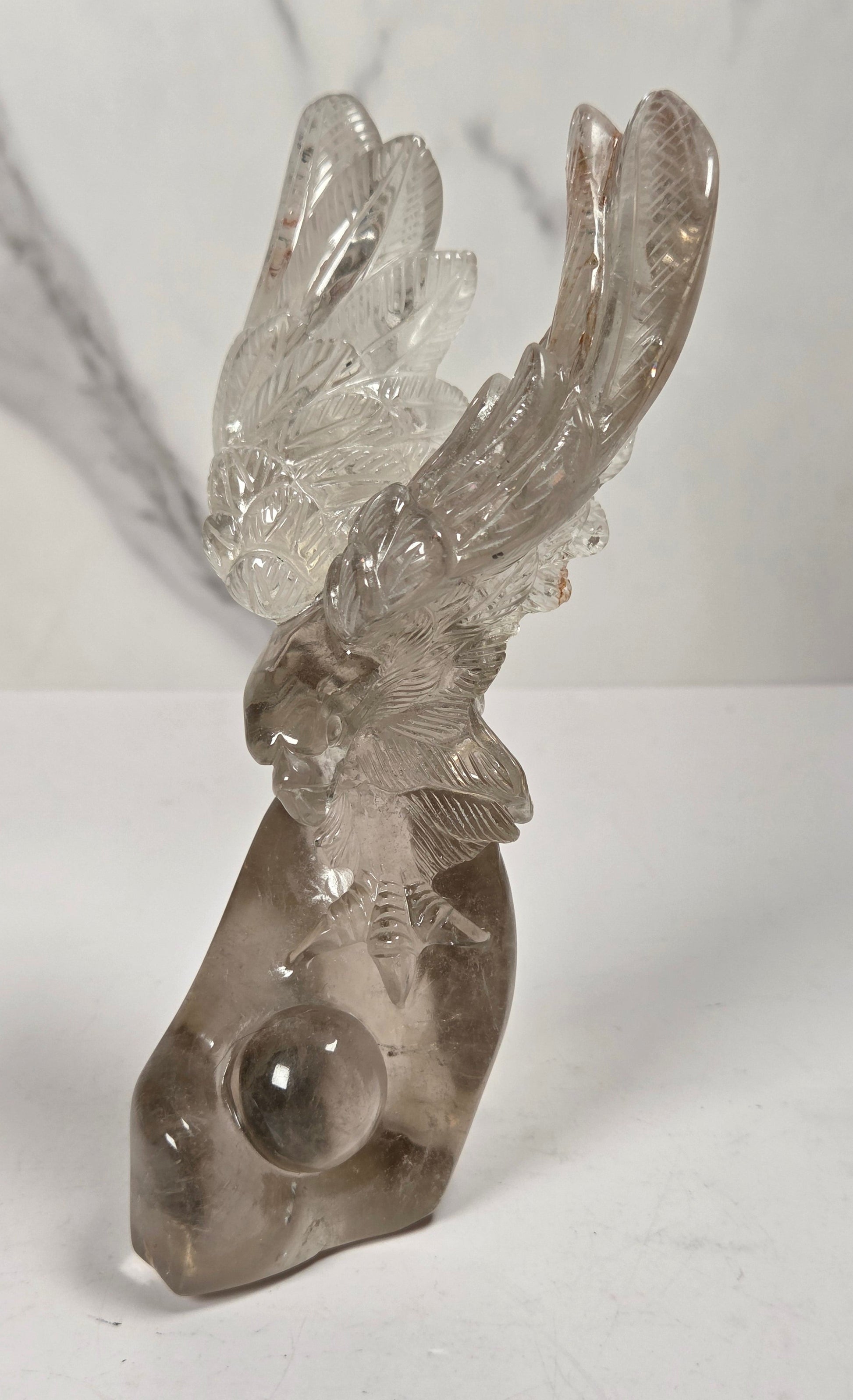 A clear smoky quartz crystal sculpture of a stag head with antlers and a round crystal ball embedded in the base, displayed on a white surface against a light gray marble-patterned background.
