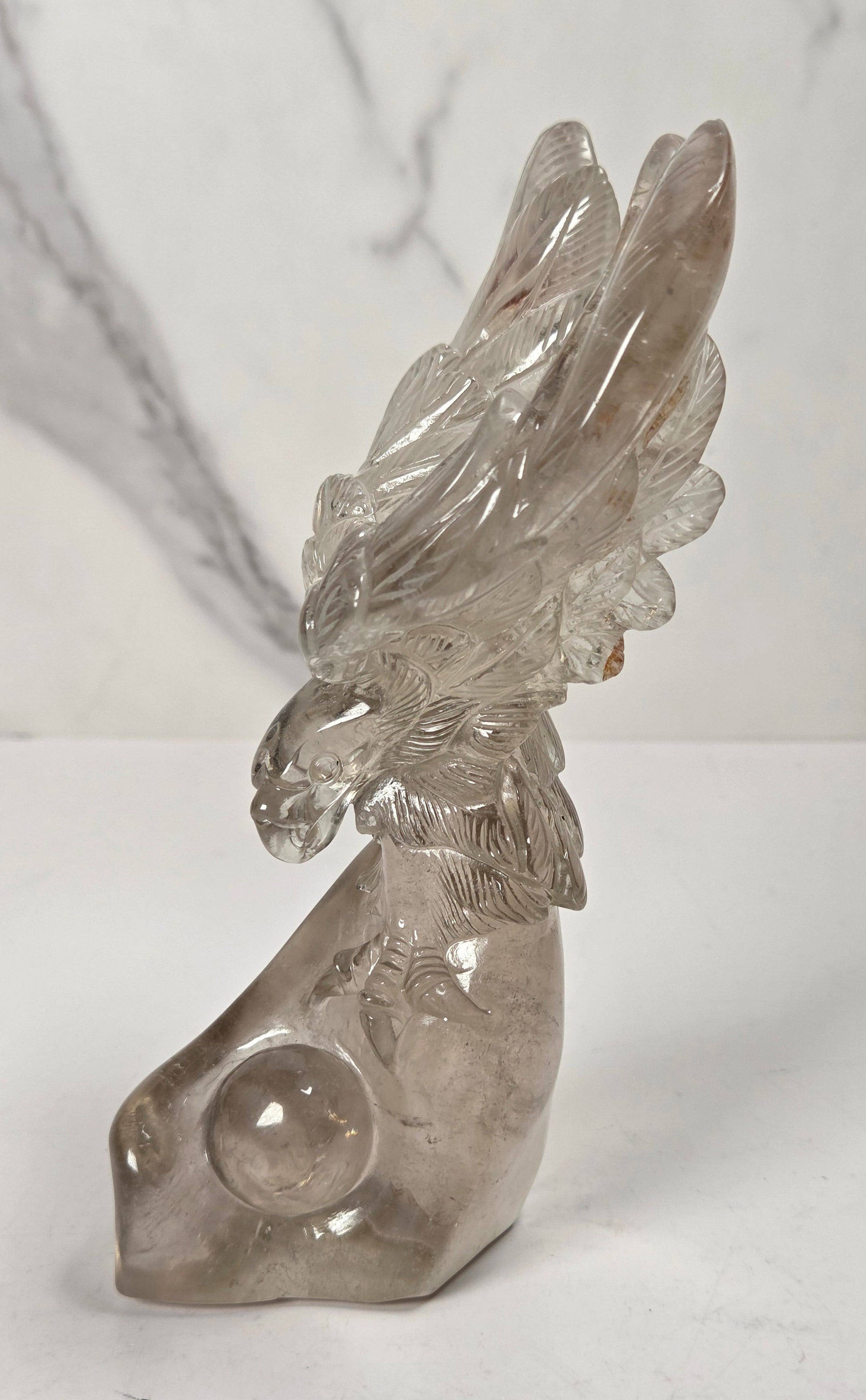 A clear quartz crystal sculpture of a bird with spread wings, displayed on a white surface against a white and gray marble-patterned background.