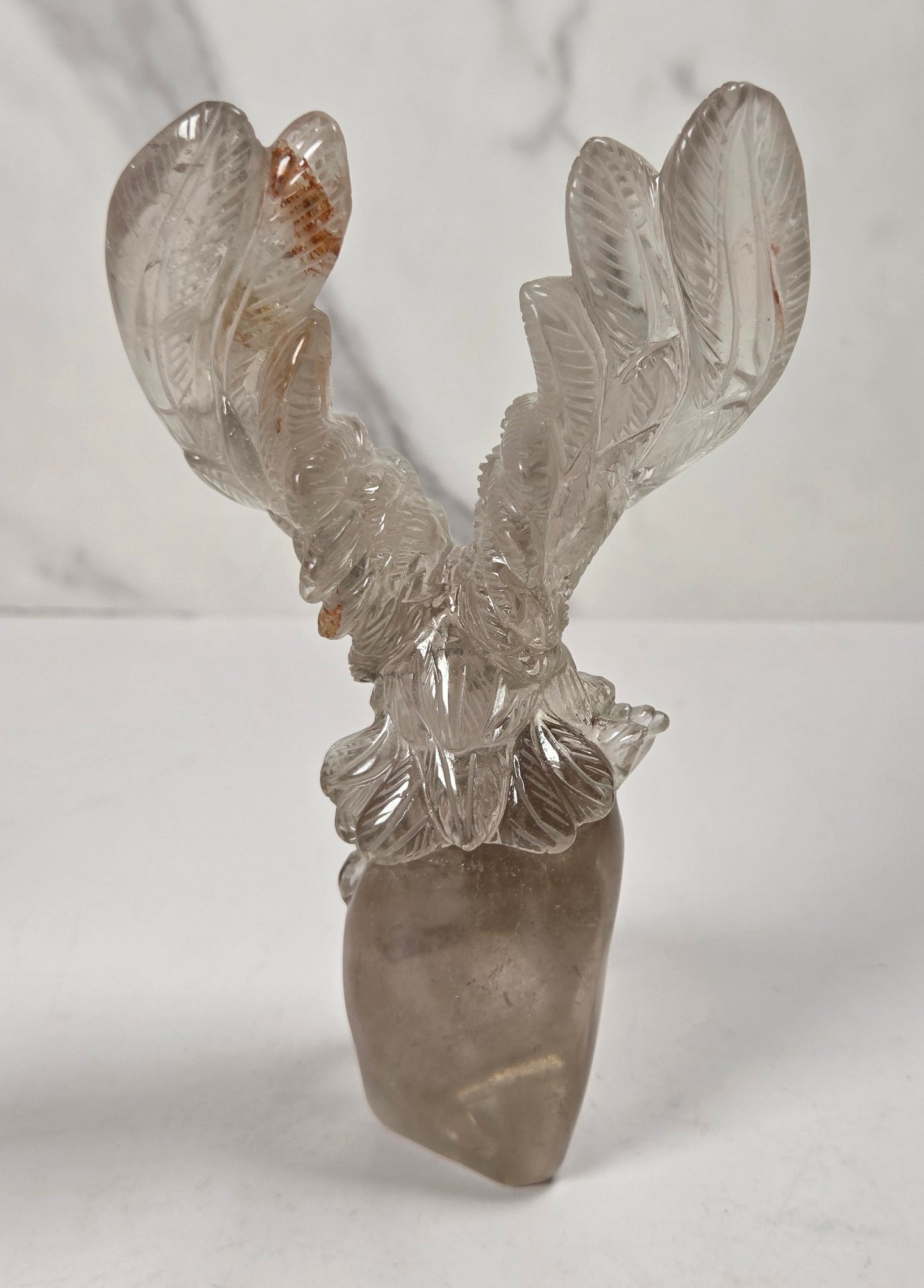 A carved crystal deer head sculpture with antlers, made from natural smoky quartz and clear quartz, displayed on a white surface against a light gray marble-patterned background.