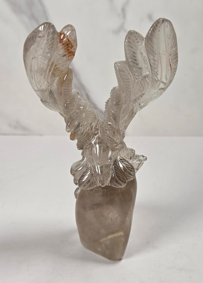 A carved crystal deer head sculpture with antlers, made from natural smoky quartz and clear quartz, displayed on a white surface against a light gray marble-patterned background.