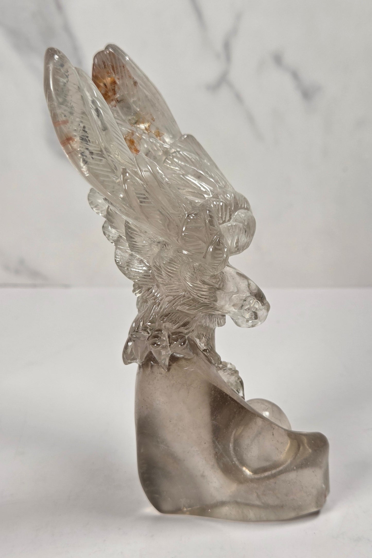 A clear glass sculpture of a stylized bird or eagle with spread wings, standing on a matching curved base against a light gray marble-patterned background.