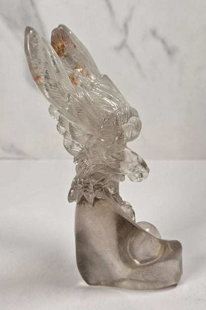 A clear glass sculpture of a stylized bird or eagle with spread wings, standing on a matching curved base against a light gray marble-patterned background.