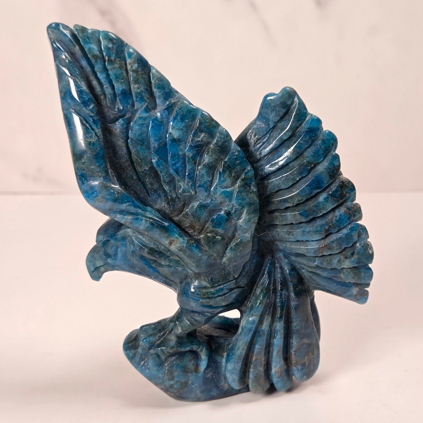 A blue stone eagle sculpture with carved wings and a textured body, displayed on a light surface against a marble-patterned background.