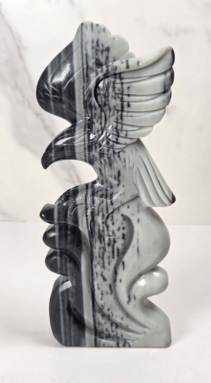 A carved stone sculpture of a stylized winged figure, likely a griffin or eagle, in polished gray and black marbled stone, standing upright on a white surface against a light marble-patterned background.